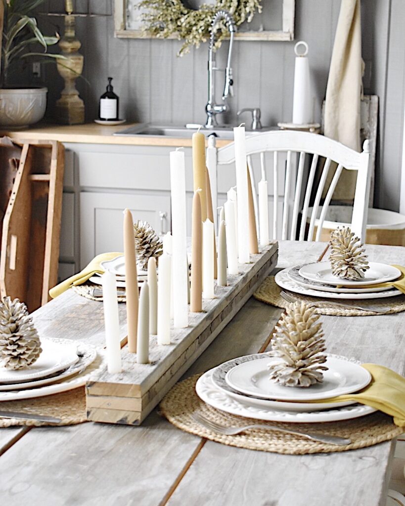 long narrow candleholder out of rustic wood with tan and gray tapers down the center of dining table with place settings of white dishes mustard color napkin on a round jute placemat with a bleached pinecone on top of each plate