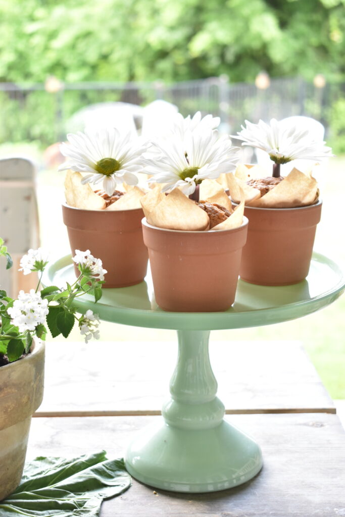 4 easy carrot muffins served in small terra cotta flower pots lined with brown tulip cupcake liners and single shasta daisy in the center or each muffin pot sitting on a green cake pedestal