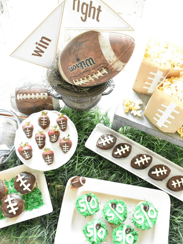 aerial view of football tailgate party dessert bar, brownie cut out of football shape cookie cutter and threads piped with white frosting, chocolate dipped strawberries with threads piped with frosting to look like footballs, cupcakes with green frosting piped with a center stripe and 50 piped on the top to resemble 50 yard line on a football field 