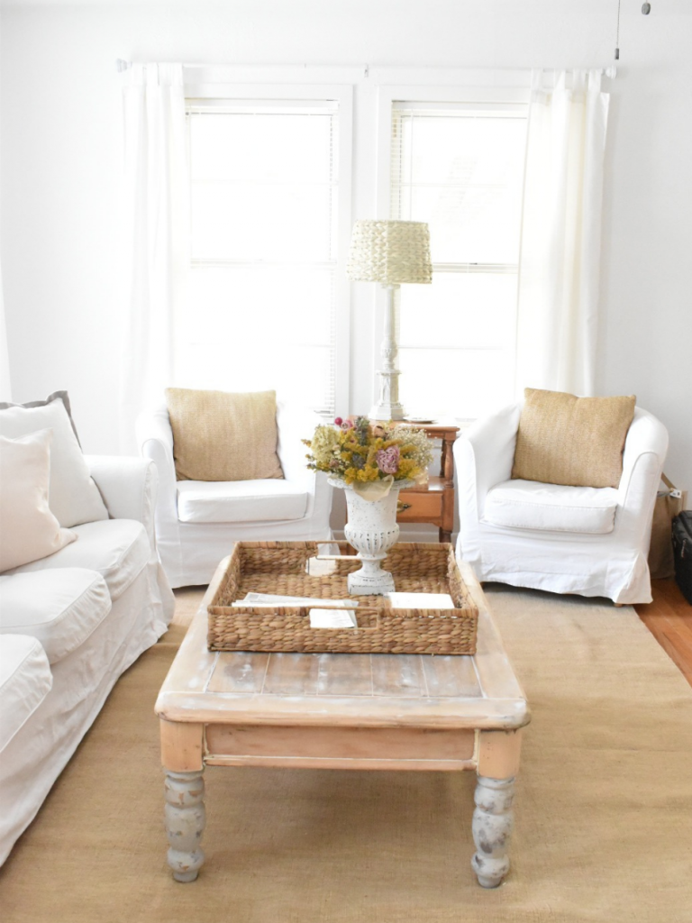 living room with 2 white slipcovered chairs white slipcovered sofa light wood coffee table light tan rug large wicker basket on coffee table with urn of flowers 