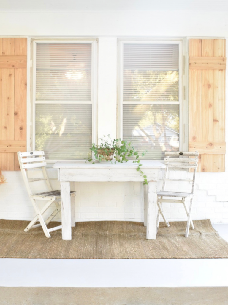 front porch of white painted brick house white table with a small white chair on either side on jute rug plant on table cedar shutters behind