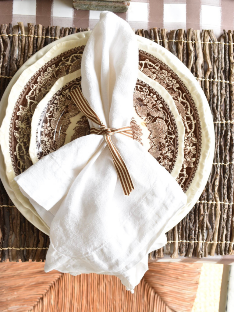 twig placemat with a large white dinner plate layered with a smaller brown transferware dinner plate topped with a white linen napkin wrapped with brown striped ribbon