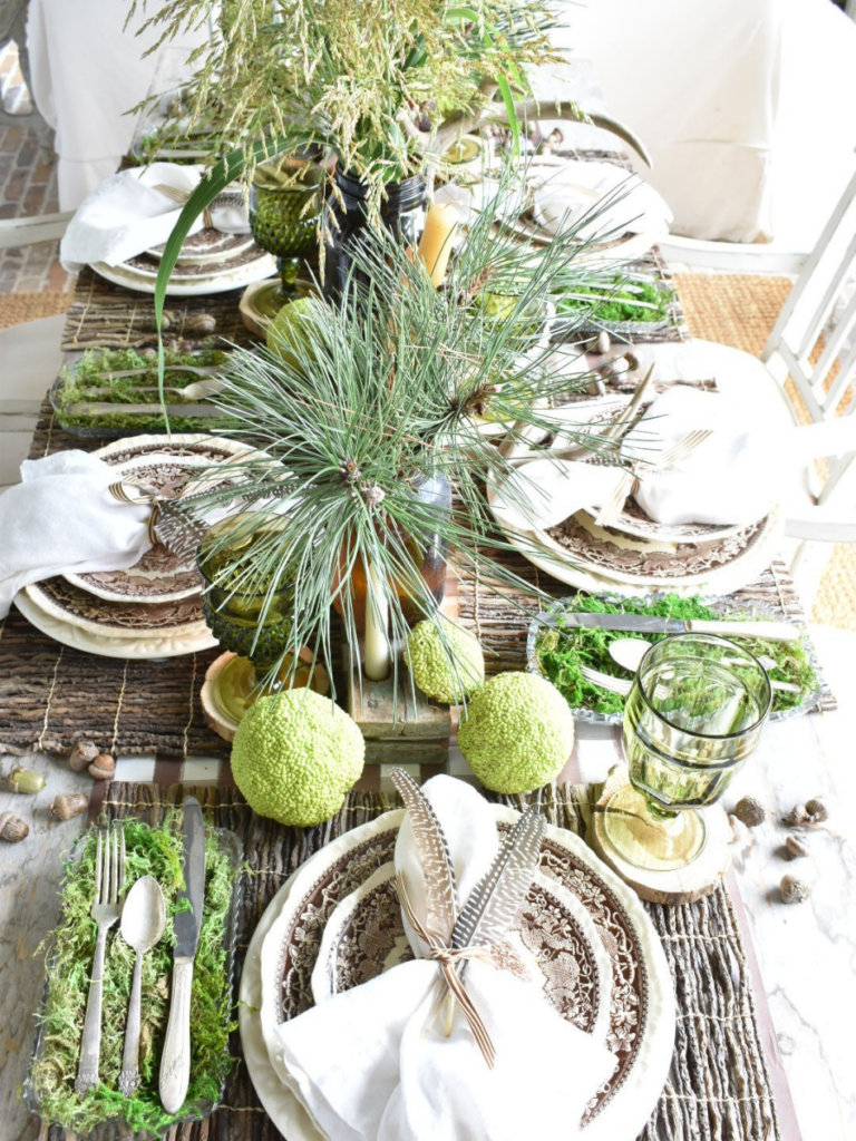 woodsy theme tablescape with brown paper plaid tablerunner twig placemat brown transferware dinner plate with brown transferware salad plate on top white linen napkin with 2 feathers on top silverware in a tray lined with green moss to the left of the place setting amber vases with pine branches for centerpiece amber bottles with tapers for centerpiece antlers acorns and horse apples for table