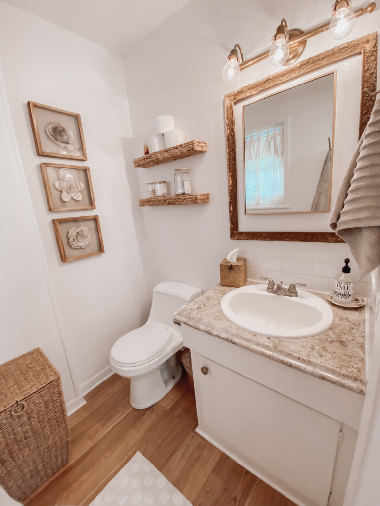 bathroom view of toilet vanity gold framed mirror woven open shelves 3 neutral flower prints wicker hamper