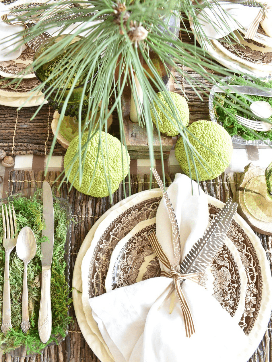 woodsy theme place setting with brown paper plaid tablerunner twig placemat brown transferware dinner plate with brown transferware salad plate on top white linen napkin with 2 feathers on top silverware in a tray lined with green moss to the left of the place setting and horse apples scattered around table for table