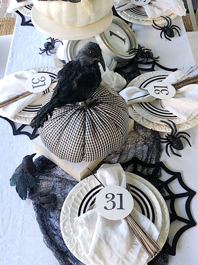 "not too spooky" halloween tablescape place setting with 2 stacked white plates white salad plate with black stripe border on top of stack white napkin on top of plate with a round card with 31 stamped on it white skeleton hand on edge of plate black felt spider web placemat black crow on top of black check fabric pumpkin