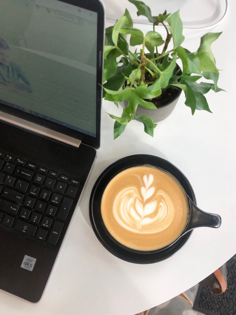 black cup with latte with plant and laptop beside it on white table