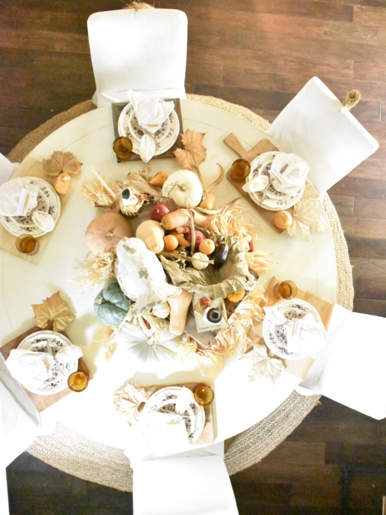 overhead view of Thanksgiving table round white dining table with thanksgiving setting wooden cutting boards for placemats white dinner plates stacked with brown transferware salad plates wooden riser in center of table with pilgrim couple dried lily pad used for cornucopia butternut squash eggplant pears mini white pumpkin spilling out of cornucopia 