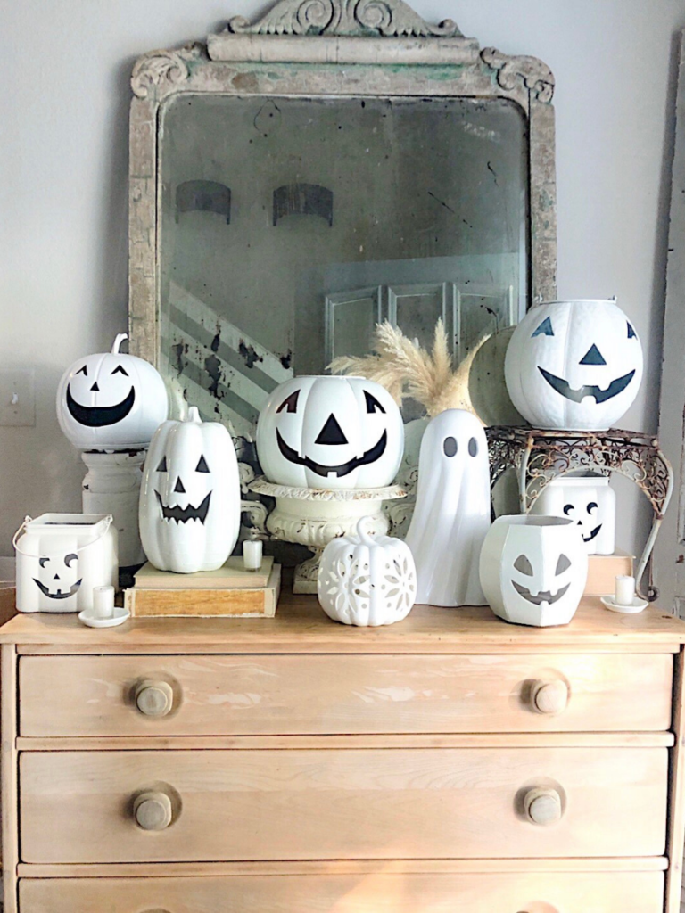 8 white jack o lanterns stacked at different heights on risers and books sitting on light wood chest and large vintage mirror in background