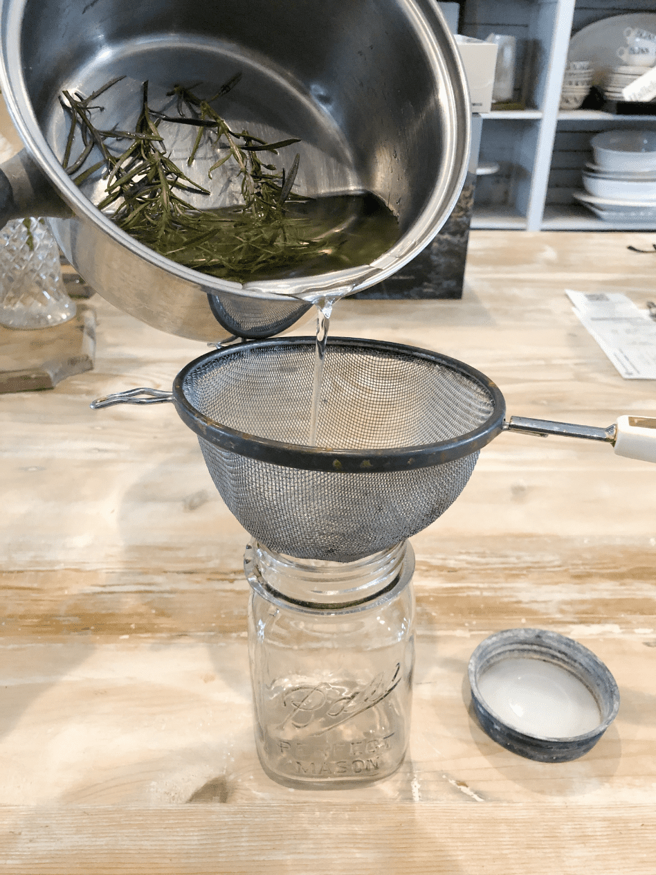 saucepan with simple syrup and rosemary sprigs pouring through strainer into a mason jar