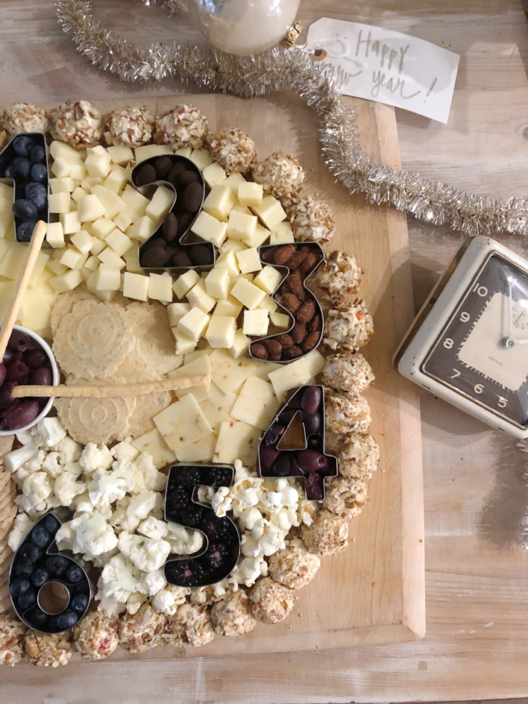 charcuterie board on a wooden cutting board in the shape of a clock with numbers 1 throught 6 white cheeses white popcorn white crackers make up the clock face and black olives blackberries blueberries and chocolate covered almonds are the number shapes