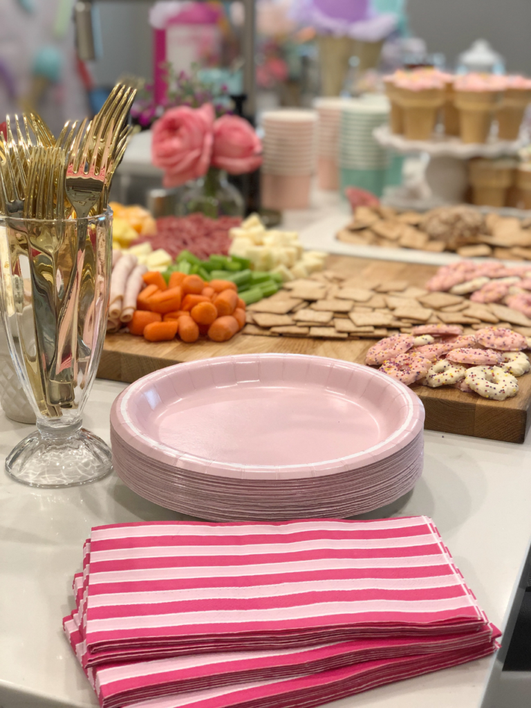 birthday party buffet table pastel pink paper plates pink and red striped napkins parfait glass with gold forks inside and charcuterie snack board in background