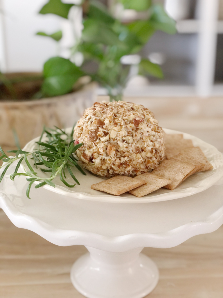 cream cheese ball with crackers and rosemary sprig on white plate on top of white pedestal and green plant in background