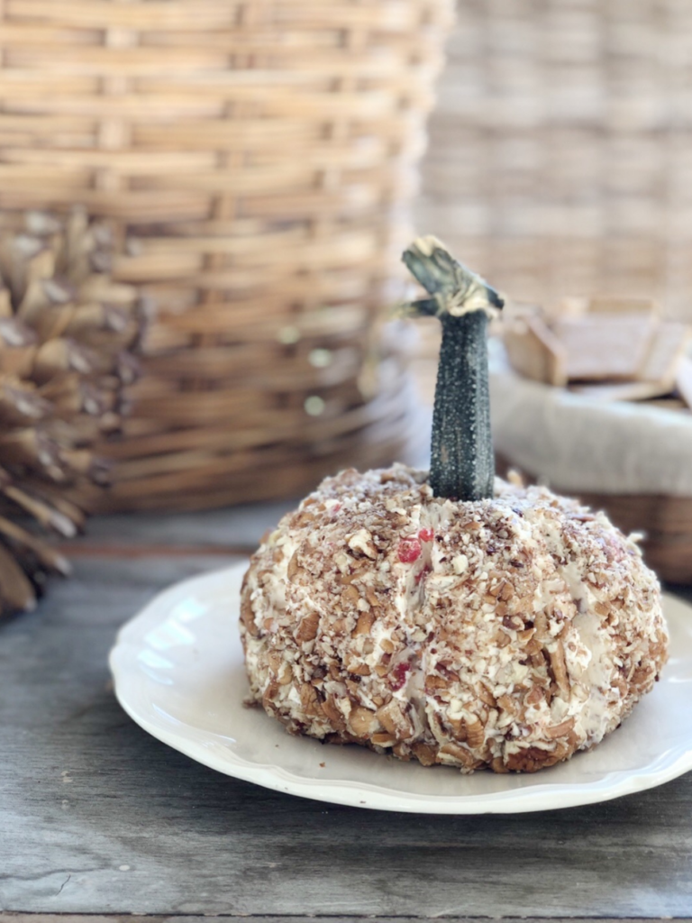 cheese ball shaped like pumpkin ridges created in sides and a real pumpkin stem on top sitting on a white plate with pinecone and wicker basket in background