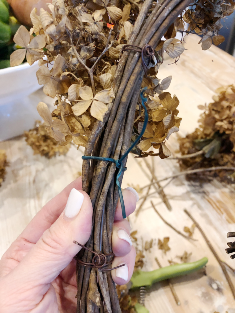 dried hydrangea bloom being tied to a grapevine wreath with a piece of wire