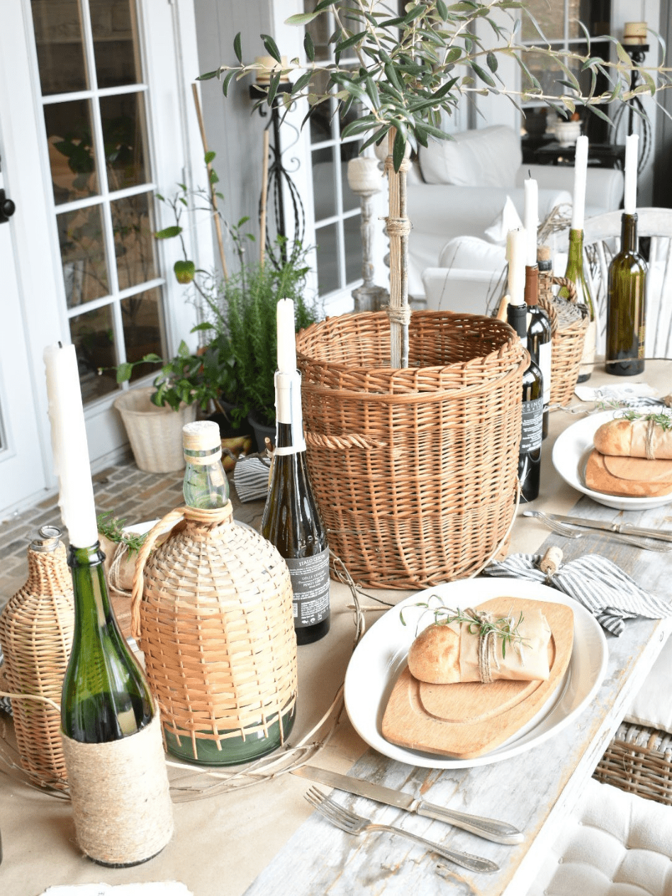 side view of rustic italian tablescape brown paper table runner down center of table 4 place settings with oval white platter oval wood board on top and a mini loaf of bread wrapped in parchment paper tied with twine and a rosemary sprig tucked in sitting on top of each plate fork and knife to left of plate and brown and white striped napkin tied with twine and a wine cork on top to right of plate wine bottles with tapers lined down center of table and a basket with a small olive tree in center of table olive oil tin with breadsticks on table grapevine woven down center of table