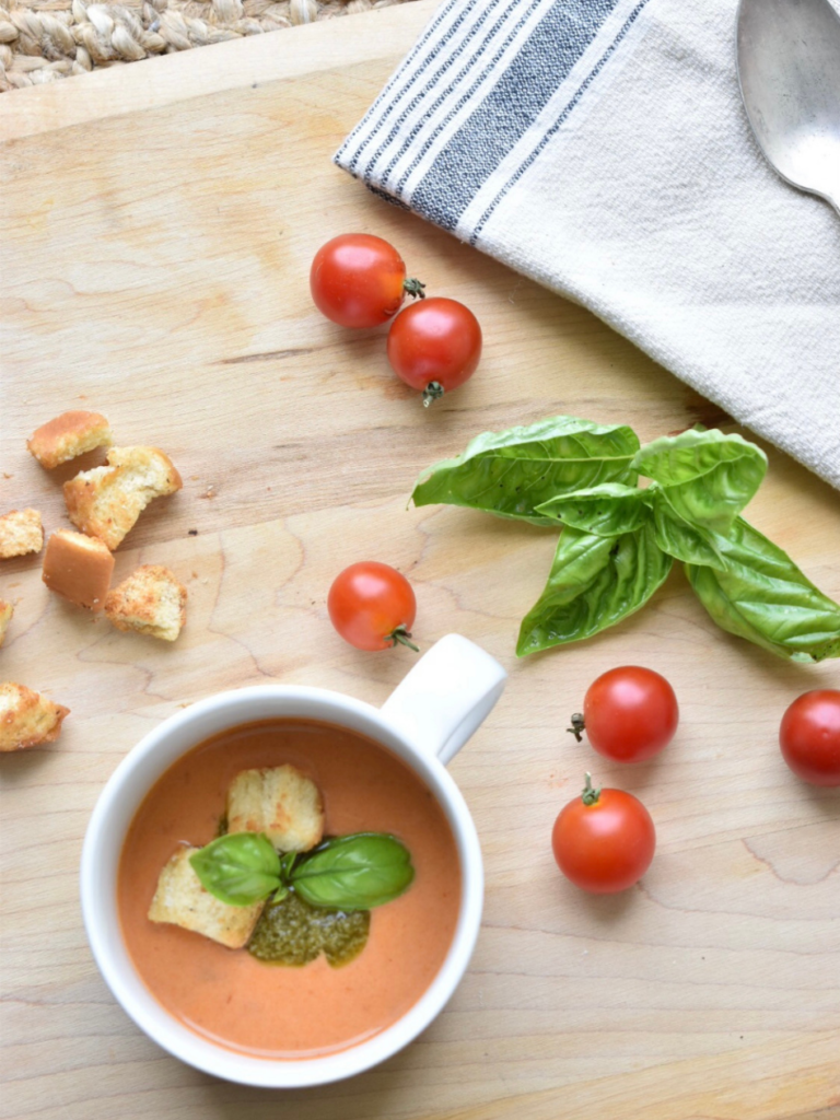 overhead view of tomato soup in a white cup with basil sprig and homemade croutons on top cherry tomatoes and basil leaves on table beside soup cup and dish towel beside