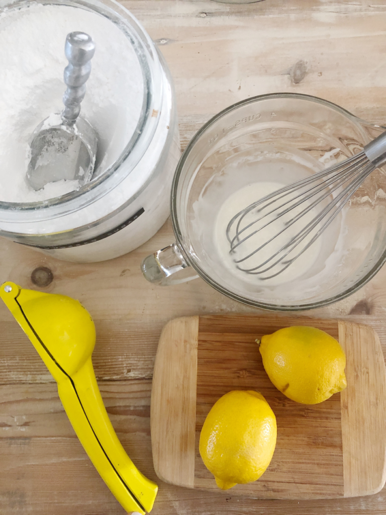 overhead view of cannister of powdered sugar with metal scoop inside 2 lemons on wooden cutting boardand lemon squeezer tool and dishtowel beside and glass mixing bowl with powdered sugar and lemon juice mixture with whisk inside bowl