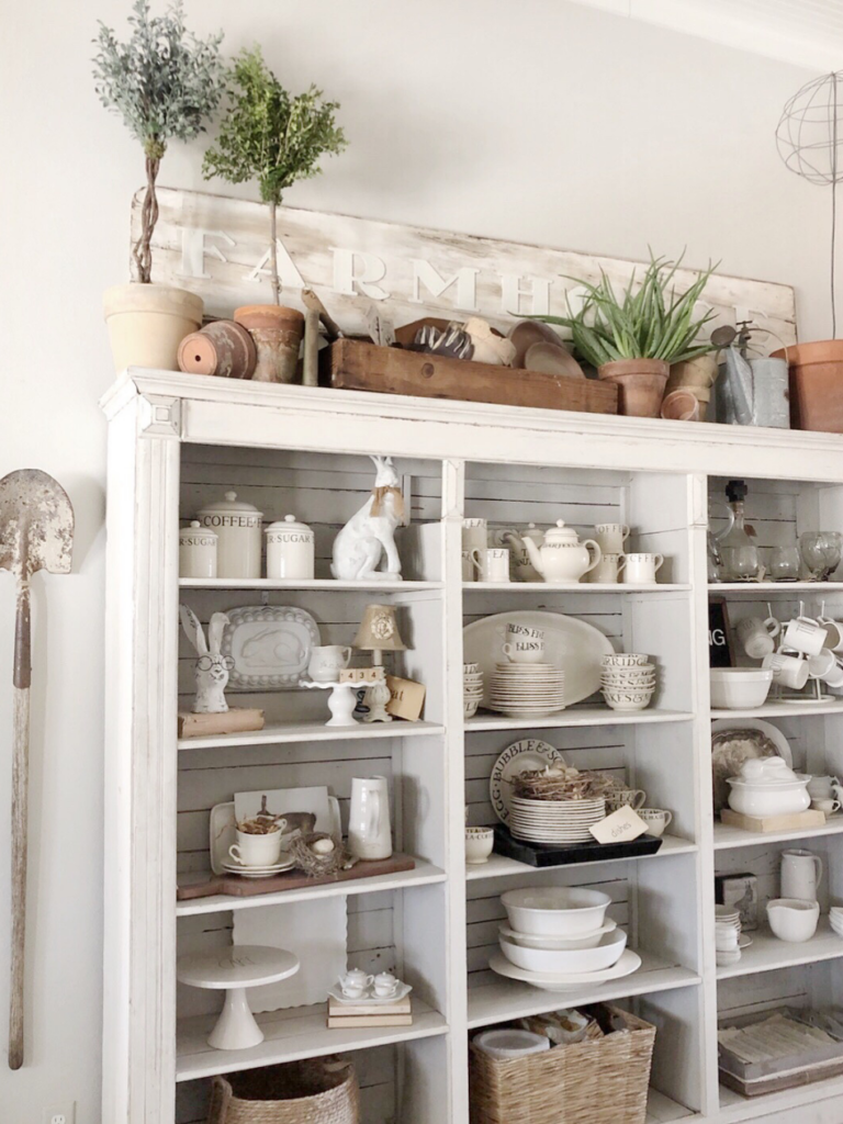 large white open front kitchen hutch with spring decor white dishes terra cotta pots plants garden tools Farmhouse sign over hutch bunnies scattered throughout dishes in hutch vintage garden shovel hanging on wall beside hutch 