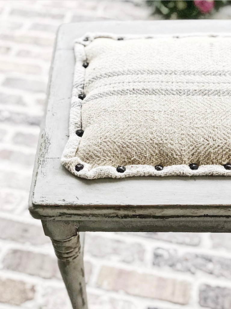 corner of a piano stool painted light gray and covered with a french grain sack with gray stripe and tacked down with nailhead studs standing on brick floor