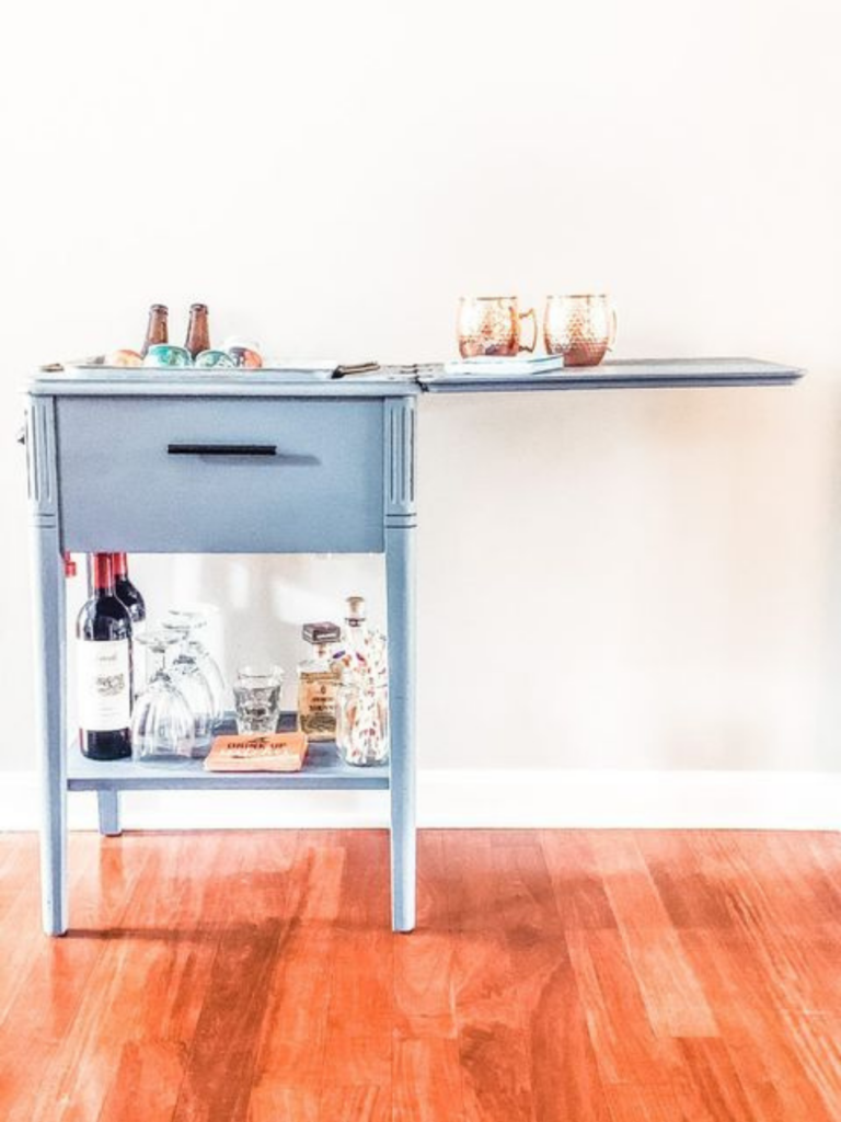 vintage sewing machine cabinet painted blue and turned into a bar cart alcohol bottles on bottom shelf drinks iced down on top of