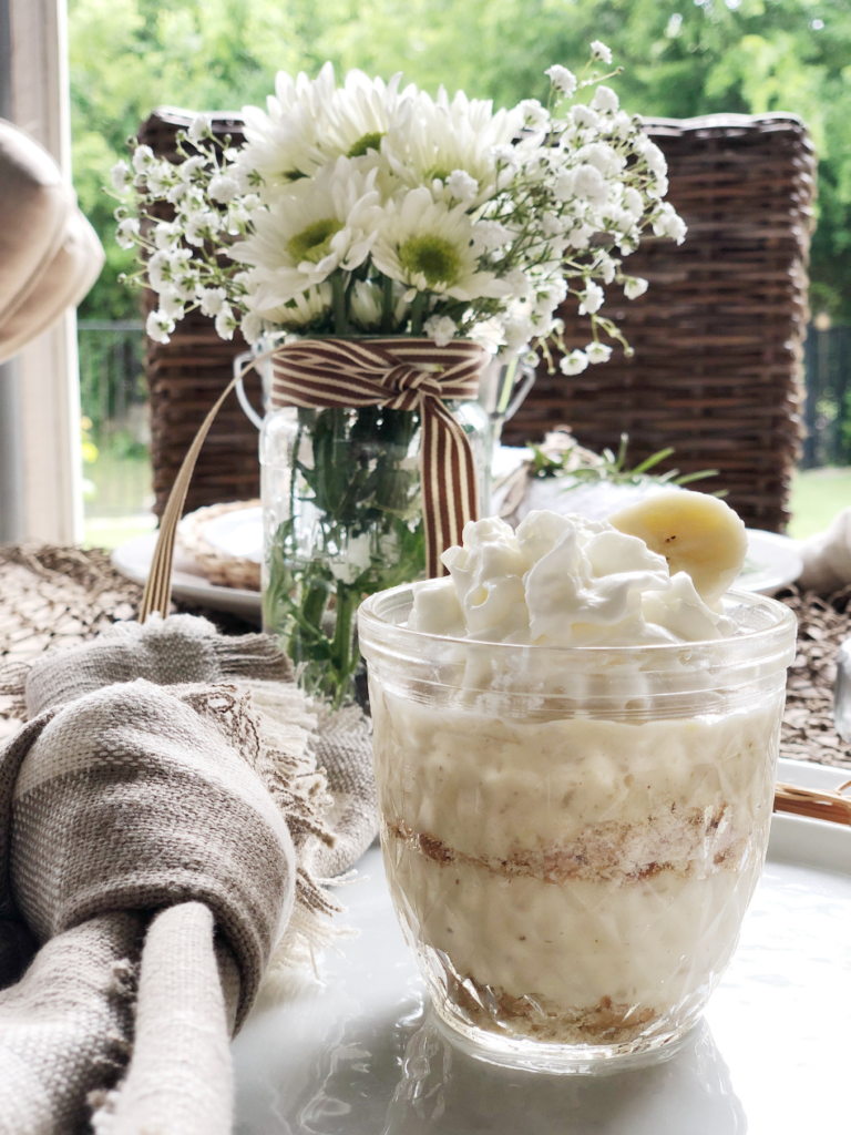 side view of small glass jar filled with banana pudding sitting on white plate with tan white check napkin beside plate mason jar of small daisies in background