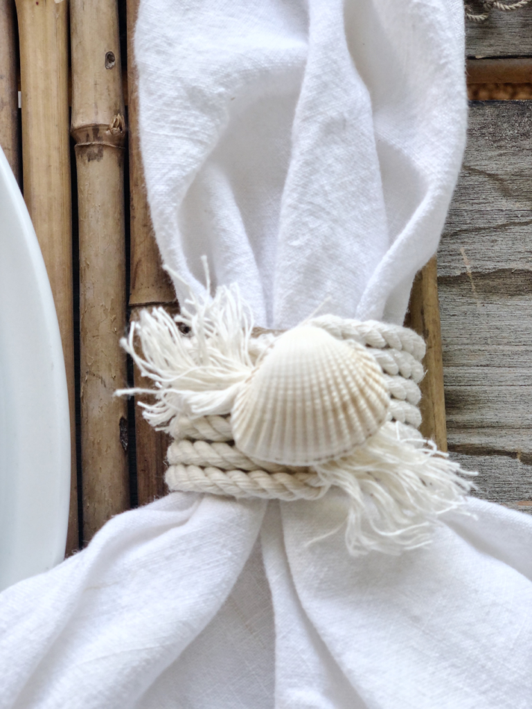 coastal napkin ring with white rope and small seashell on top of rope with white napkin pulled through napkin ring sitting on bamboo placemat