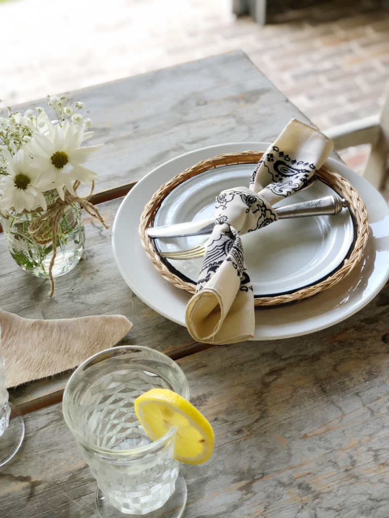 side view of simple backyard barbeque place setting white dinner plate with wicker plate holder on top and white enamelware plate inside tan/black bandana tied around fork and knife sitting on top of plate mason jar of baby's breath and daisies to right of plate clear glass of water with lemon wedge on side to left of plate