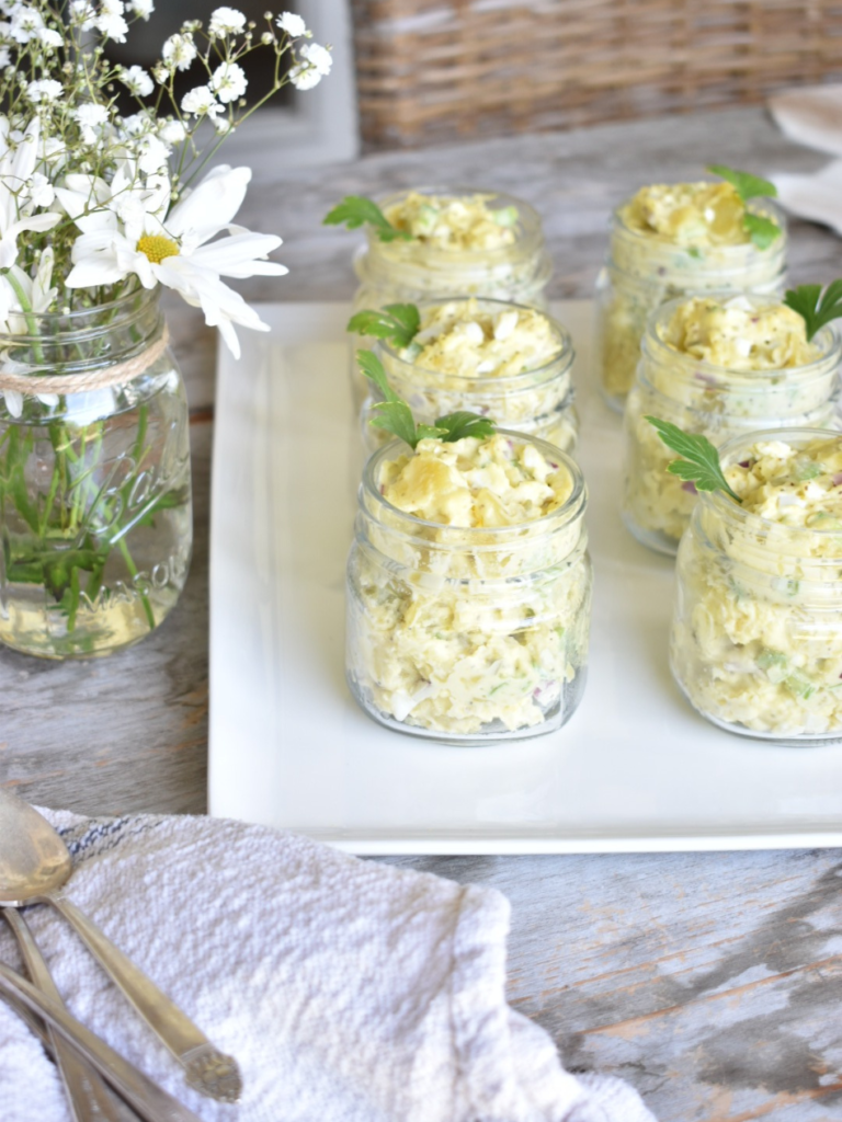 6 small mason jars filled with heavenly potato salad sitting on white tray garnished with a parsley leaf and vase of white daisies beside tray