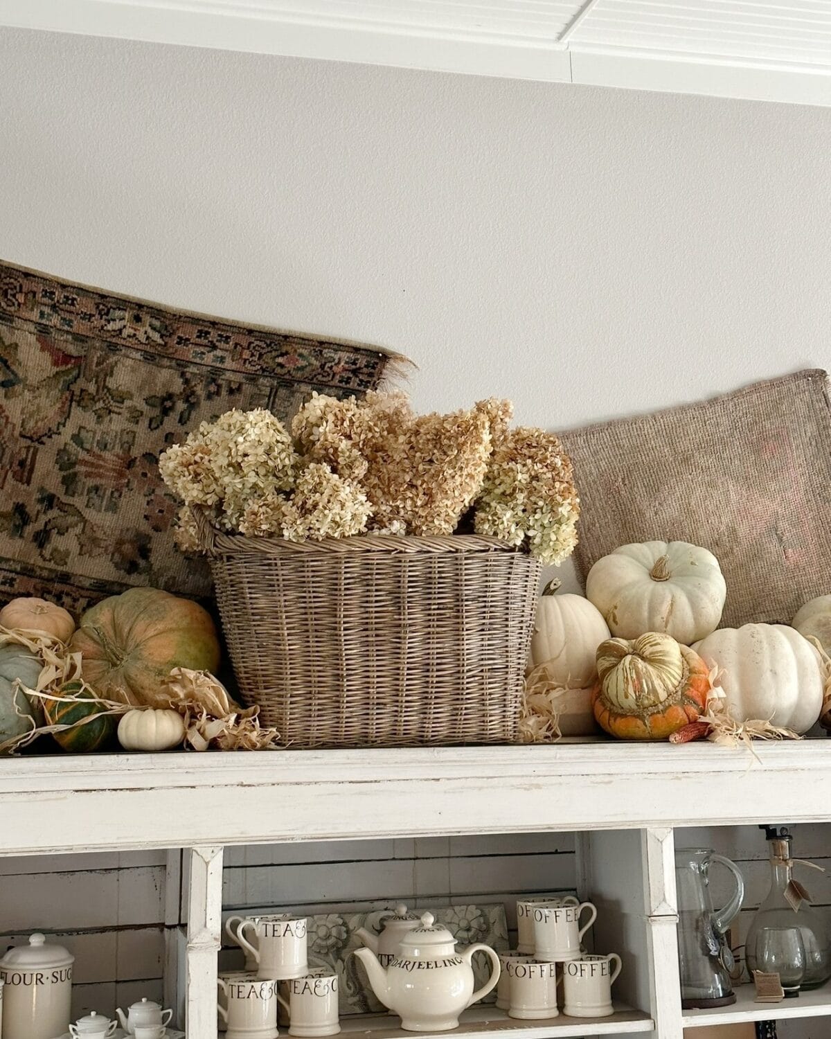 A woven basket filled with dried hydrangeas sits on a shelf, surrounded by assorted white and green pumpkins. Decorate With Dried Hydrangeas For Fall alongside vintage teapots, cups, and antique textiles for a charming seasonal display.