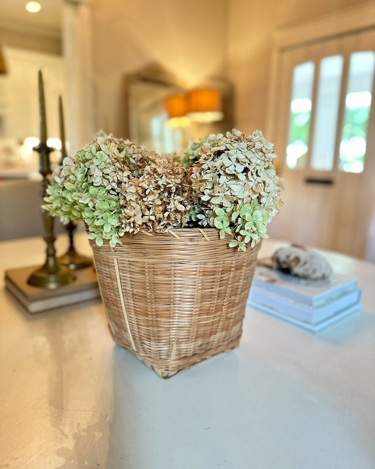 A wicker basket filled with light green and dried hydrangeas sits on a white table. In the background, candlesticks, stacked books, a shell, and a mirror reflecting a lit lamp show how to Decorate With Dried Hydrangeas For Fall.