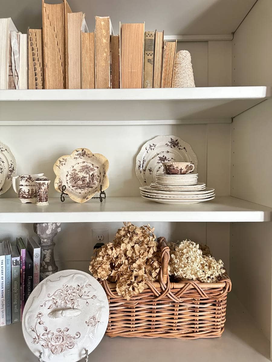 A bookshelf holds neutral-toned books, vintage china plates and cups with floral patterns, a basket filled with dried hydrangeas to Decorate With Dried Hydrangeas For Fall, and a large decorative plate with a floral design on the lower shelf.