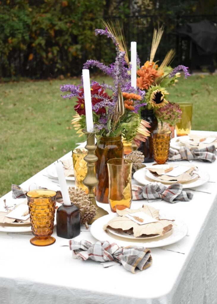 fall tablescape on long table outside amber glasses white dinner plates with large leaf on top of plate amber vases filled with fall color flowers