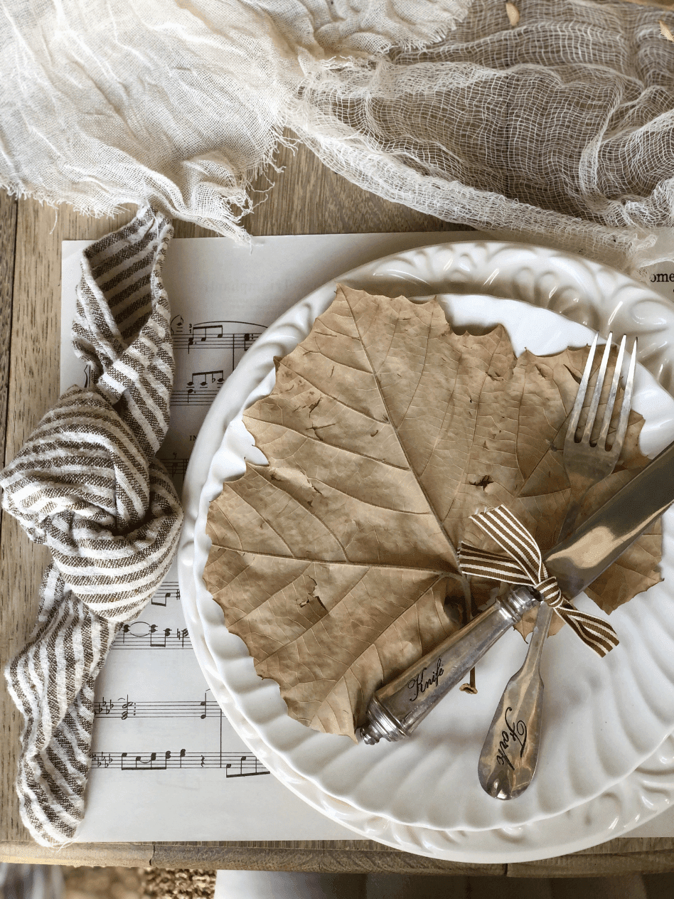 place setting for a simple fall tablescape sheet music placemat white dinner plate layered with white salad plate large dried leaf on plate silverware tied with striped ribbon on plate