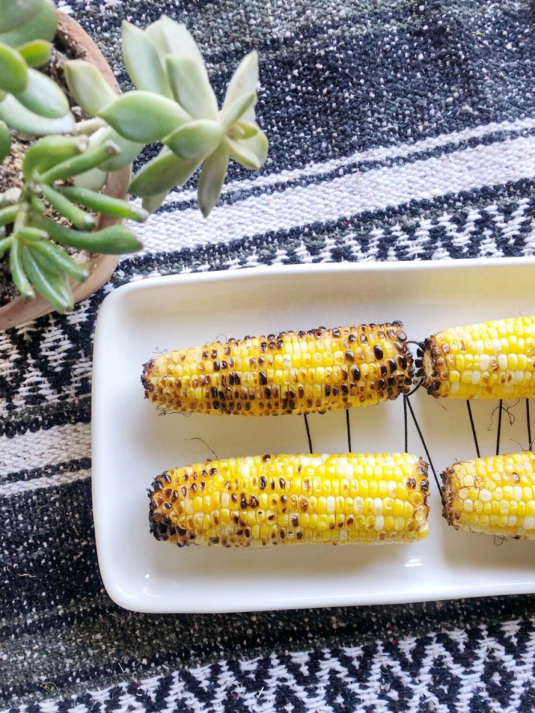 A white rectangular plate with four grilled corn cobs arranged in a row sits on a patterned black-and-white tablecloth. Adjacent to the plate is a potted succulent plant with green, fleshy leaves spilling over the edge of its pot.