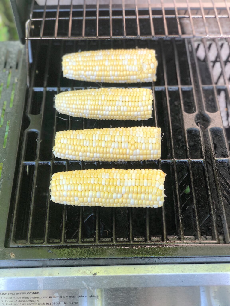 Four ears of corn with husks removed are arranged in a single layer on a metal grill rack, ready to add a smoky depth to your black bean and corn nachos. The grill appears to be turned on, and the surrounding area is clean with some grill marks visible on the rack. The view is a close-up from above.