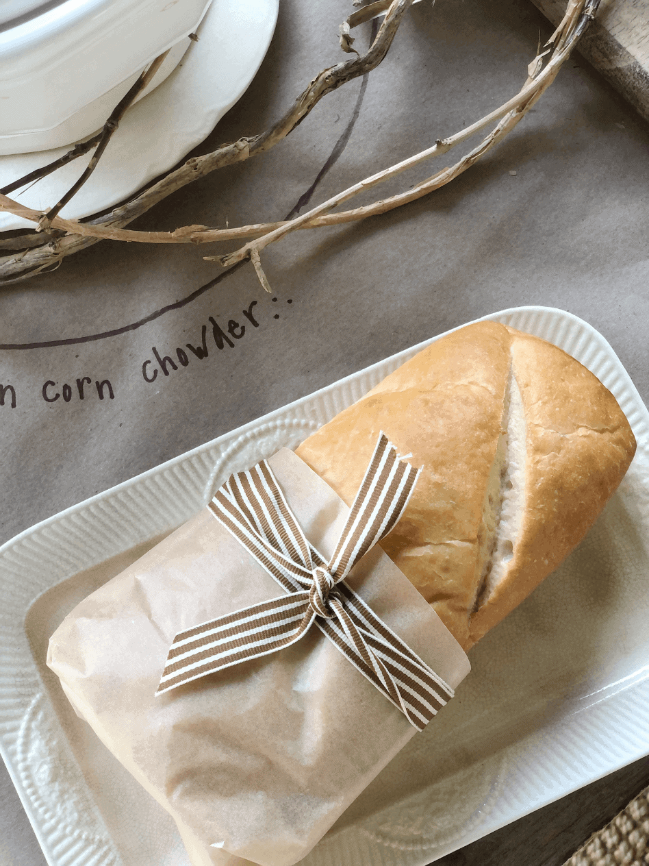 french bread wrapped with brown parchment tied with brown strip ribbon sitting on white platter