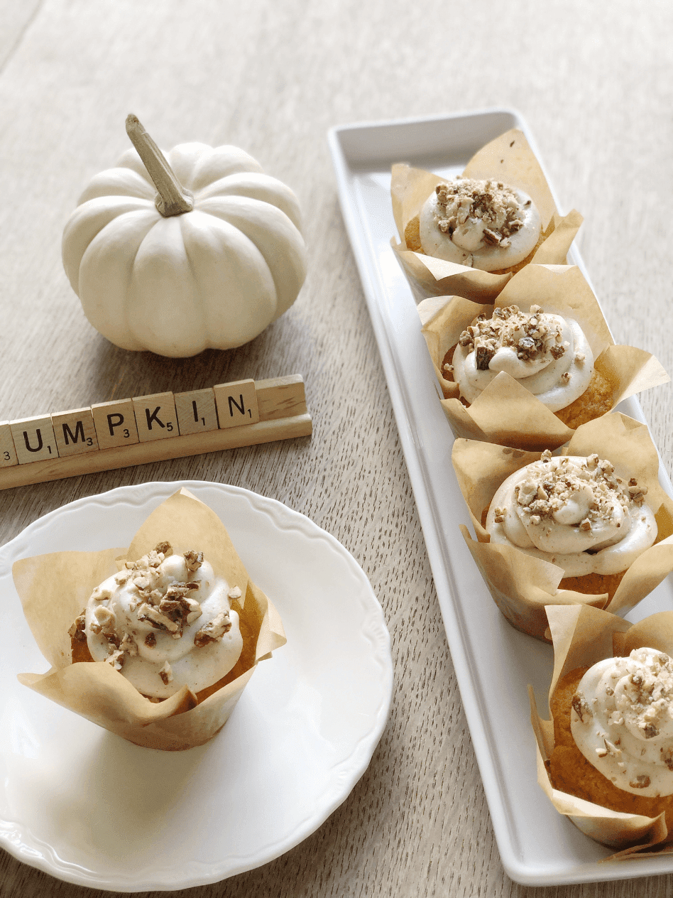 long white tray with 5 pumpkin cupcakes with cream cheese frosting topped with sugared pecans white dessert plate with 1 cupcake scrabble tiles "pumpkin" mini white pumpkin