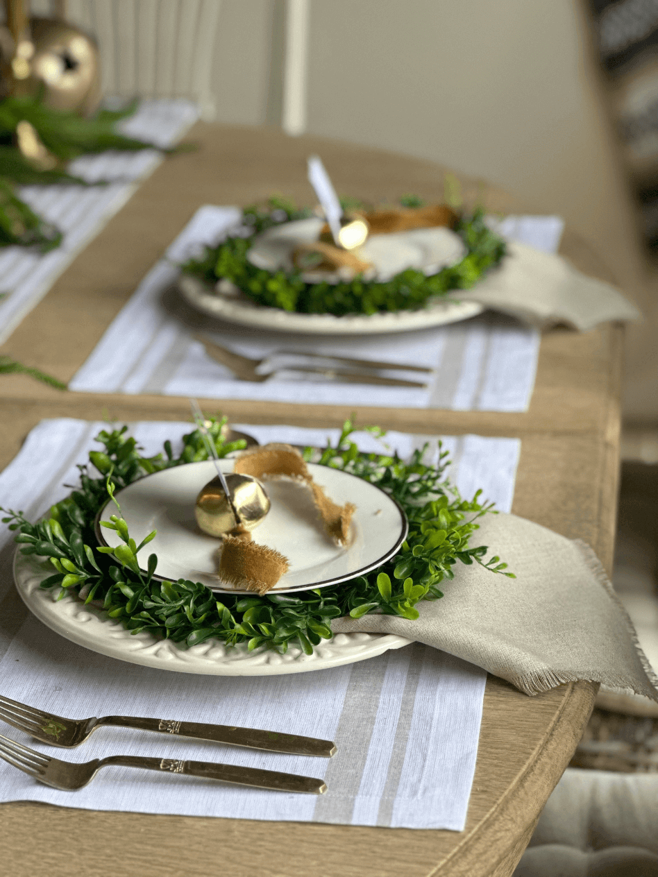 2 christmas place settings white linen placemat gold flatware white dinner plate small greenery wreath with white salad plate on top gold bell with gold felt ribbon on top of plate for place card holder linen napkin under dinner plate