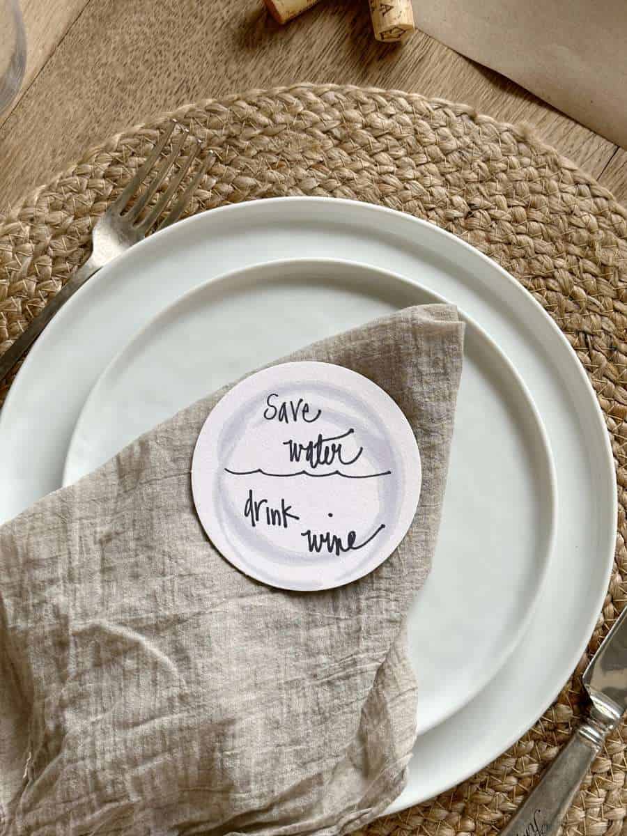 place setting for wine and cheese gathering jute placemat white plate linen napkin on top of plate with wine saying coaster on top