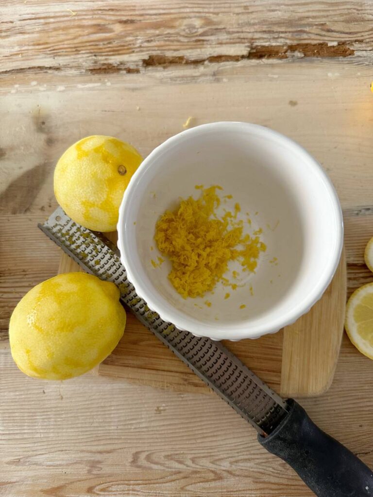 white bowl with lemon zest 2 lemons beside bowl and lemon zester