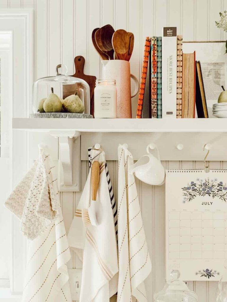 white wall shelf with cookbooks on shelf cloche with pears under dishtowels hanging on pegs