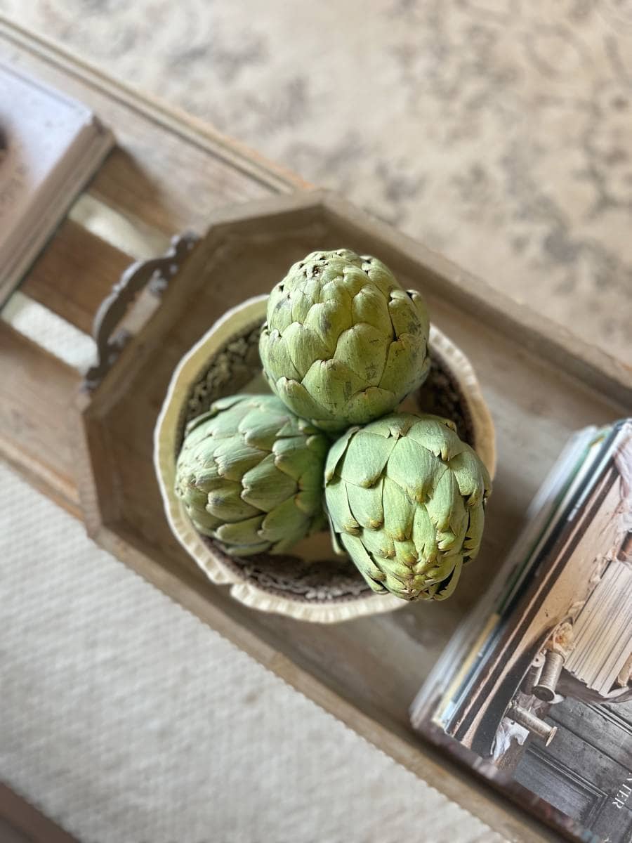 A top-down view of a wooden tray holding a bowl with three artichokes. Next to the tray, there is a stack of books on a light-colored, textured surface, possibly a carpet or rug. The overall setting appears rustic and cozy, perfectly capturing the change from summer to fall on a budget.