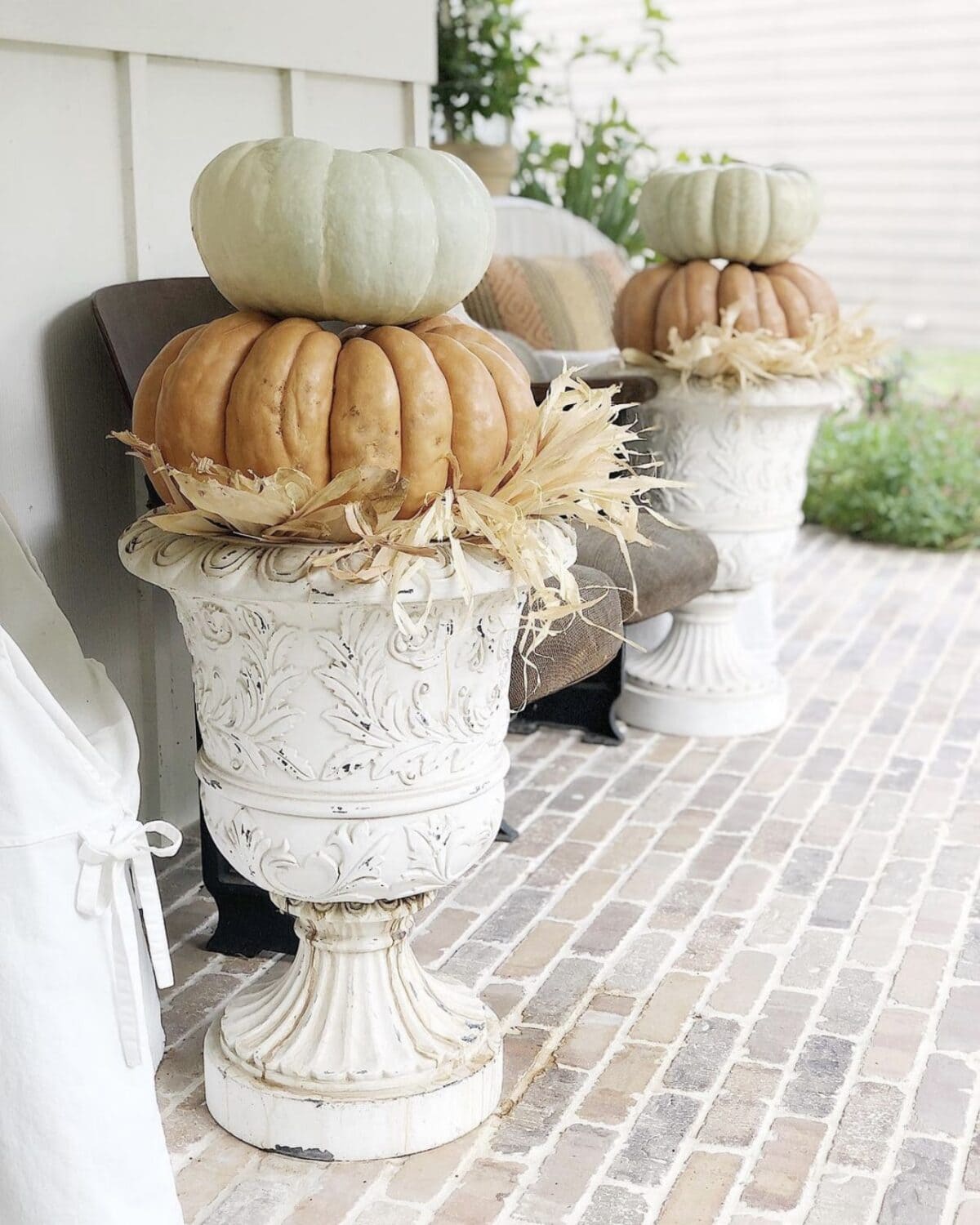 Two white ornate urn planters on a brick porch hold a stacked arrangement of pumpkins, showcasing how to change from summer to fall on a budget. Each planter features a large orange pumpkin with a smaller green pumpkin on top, accented with dried corn husks around the base. Brown chairs are visible in the background.