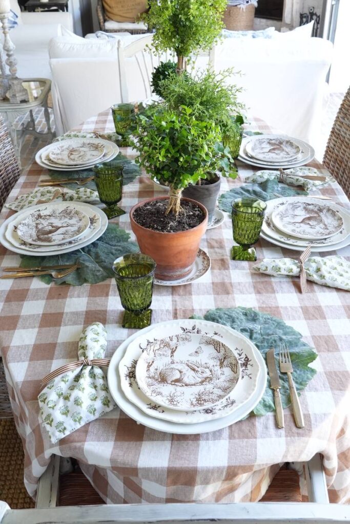 brown check tablecloth with brown pattern bunny plate green block print napkin gold flatware on cabbage leaf placemat at 6 place settings for spring table topiaries down center of table