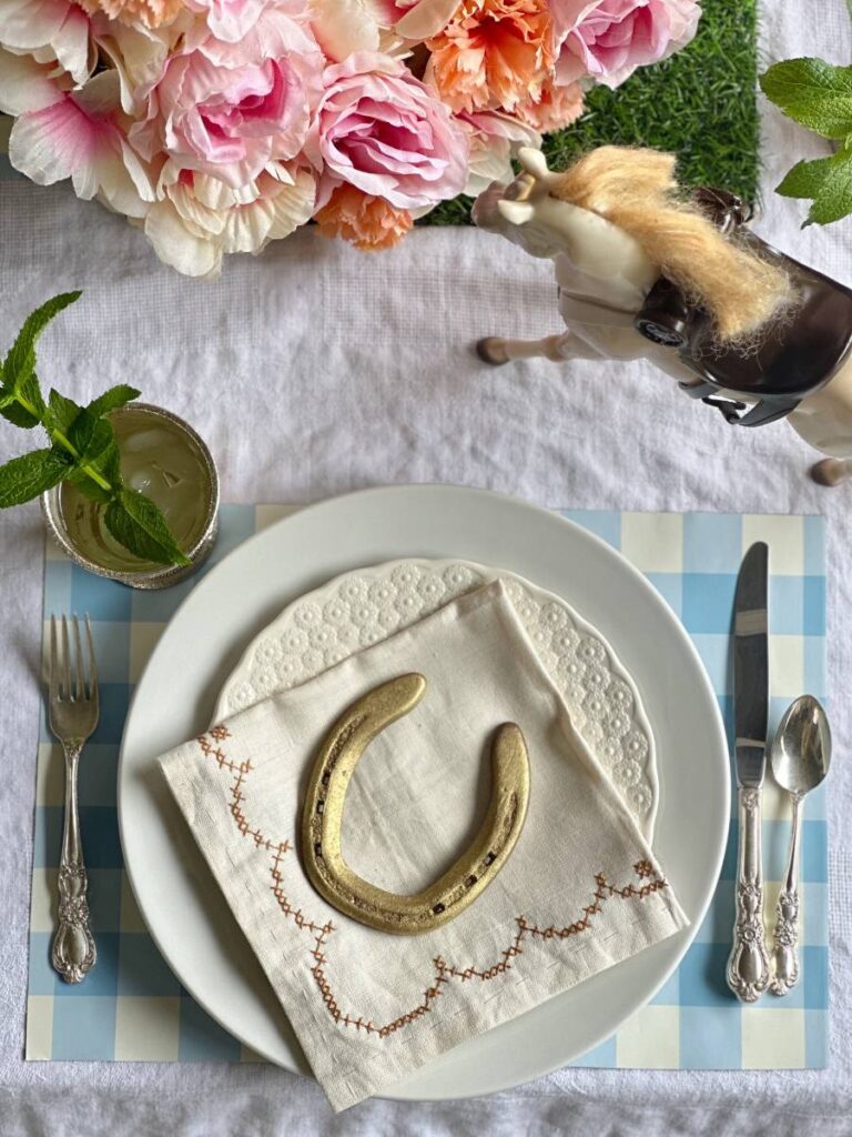 white dinner plate stacked with white salad plate folded napkin on top and gold horseshoe on blue and white check placemat and mint julep cup beside plate for Kentucky Derby place setting