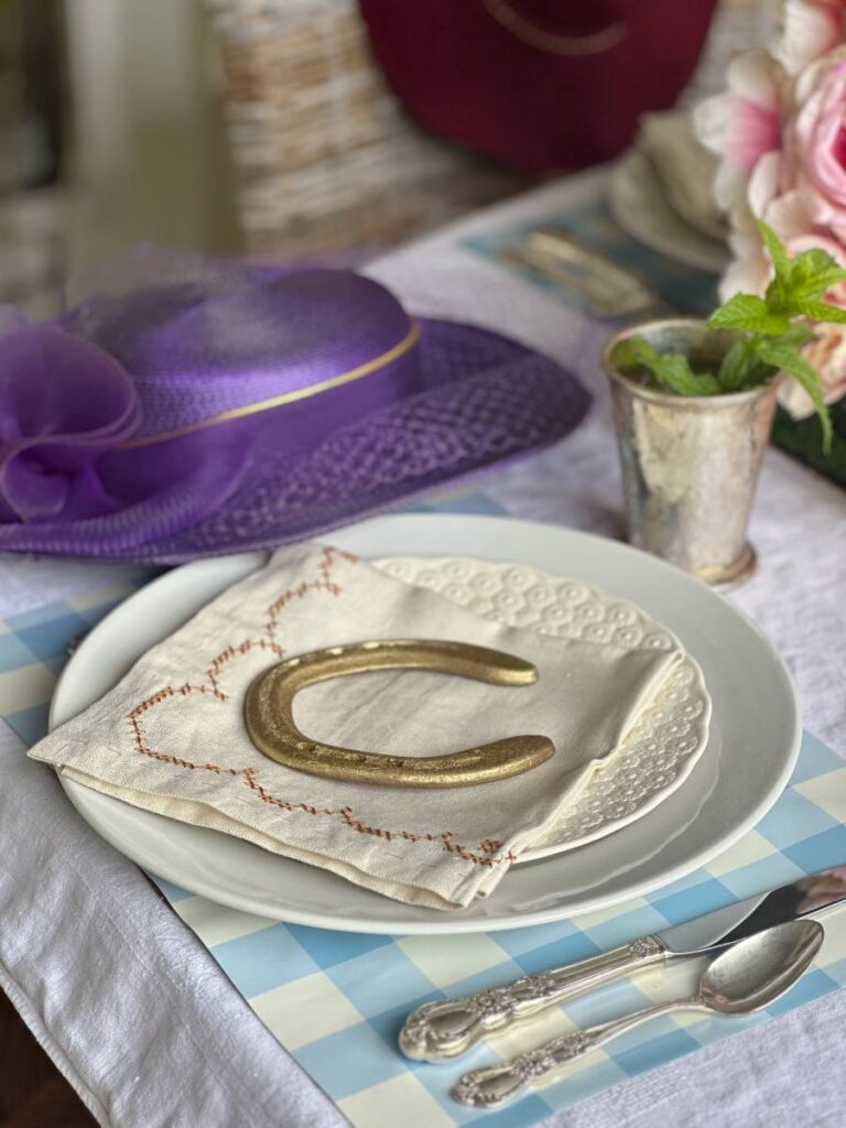 white dinner plate stacked with white salad plate folded napkin on top and gold horseshoe on blue and white check placemat and mint julep cup beside plate for Kentucky Derby place setting