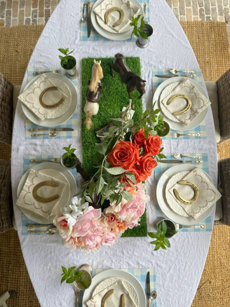 budget friendly Kentucky Derby table setting with white dinner plate stacked with white salad plate folded napkin on top and gold horseshoe on blue and white check placemat and mint julep cup beside each plate strip of turf down center of table with 3 toy horses cup trophy with roses horse trophy for centerpiece