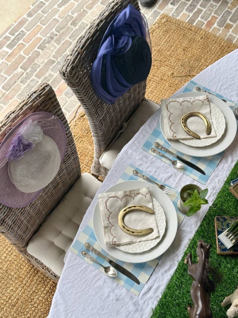 2 place settings of white dinner plate stacked with white salad plate folded napkin on top and gold horseshoe on blue and white check placemat and mint julep cup beside plate for Kentucky Derby dinner table fancy hats on back of chairs