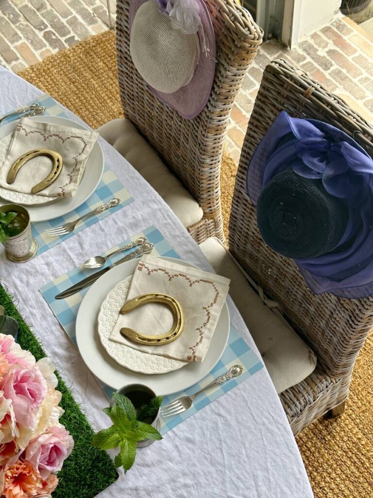 2 place settings of white dinner plate stacked with white salad plate folded napkin on top and gold horseshoe on blue and white check placemat and mint julep cup beside plate for Kentucky Derby dinner table fancy hats on back of chairs